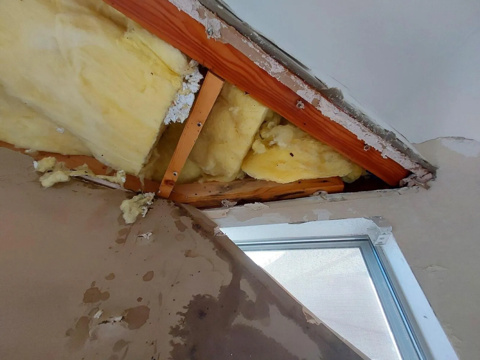 A ceiling opened above a window, with yellow insulation and wood framing showing through the gap and dark water stains running down the tan wall below