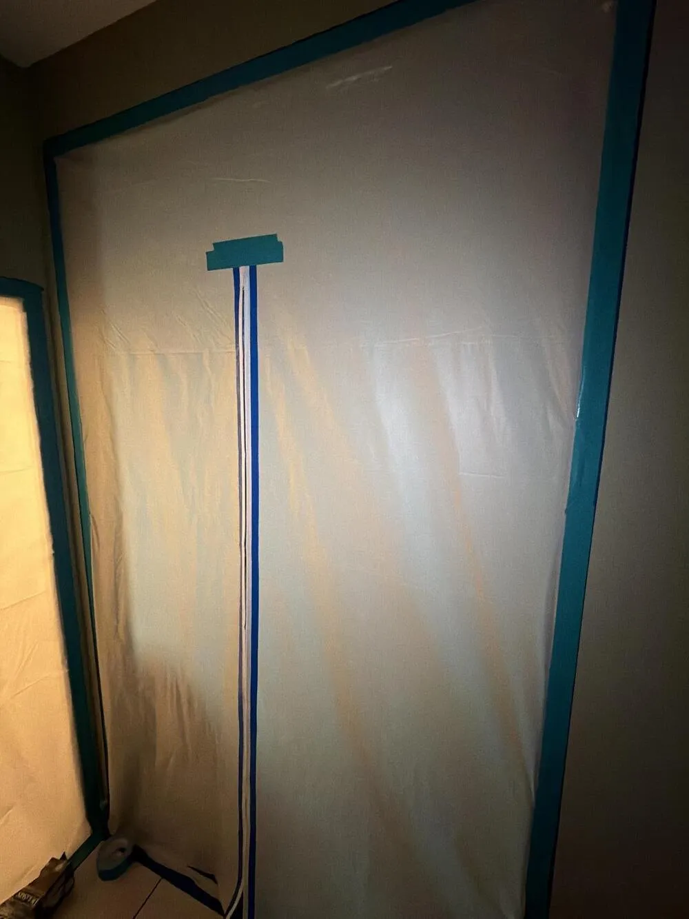 Translucent white plastic sheeting sealed to a wall with teal tape, with a zipper door edged in blue tape running down it left of center, beside a second plastic-covered opening lit from behind