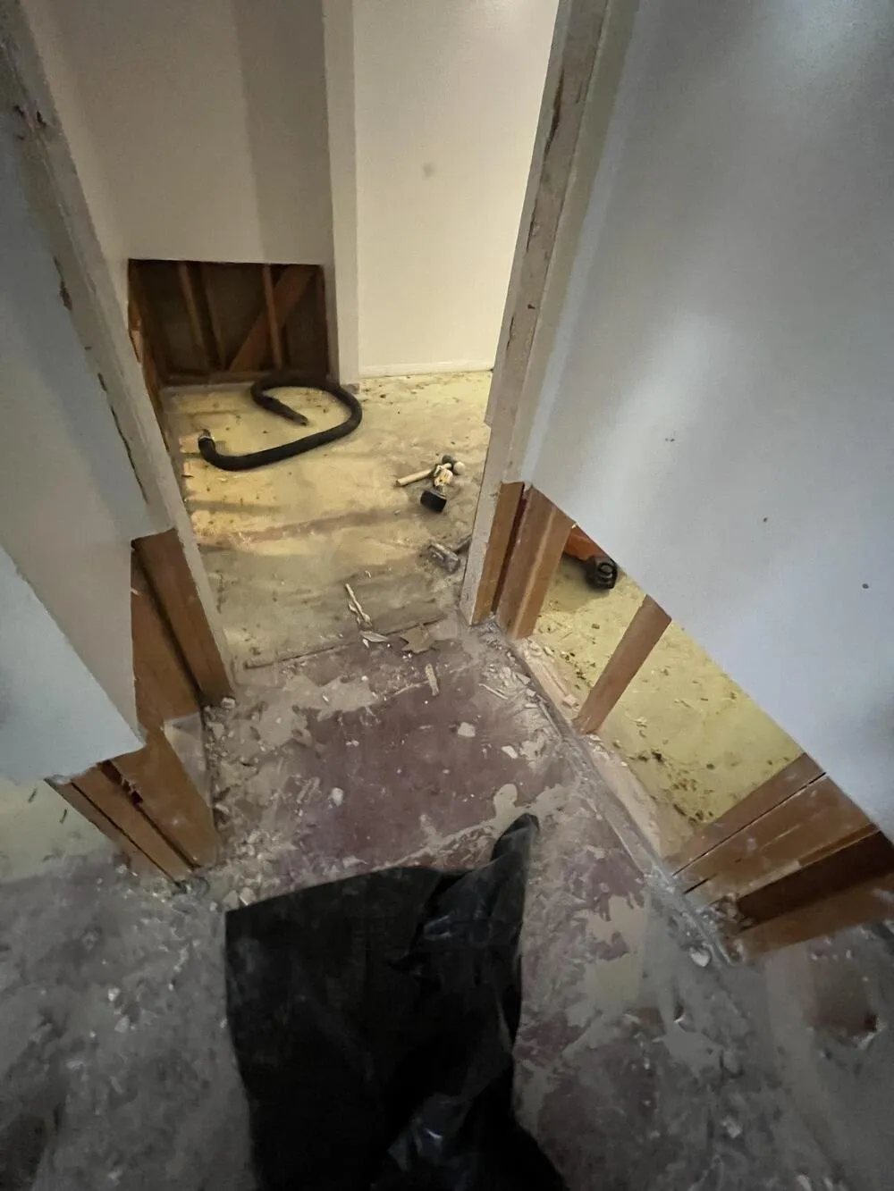 A hallway with the bottom of the drywall cut away to the wood studs and the flooring pulled up to a bare, dusty floor, with a black hose and hand tools left on the floor