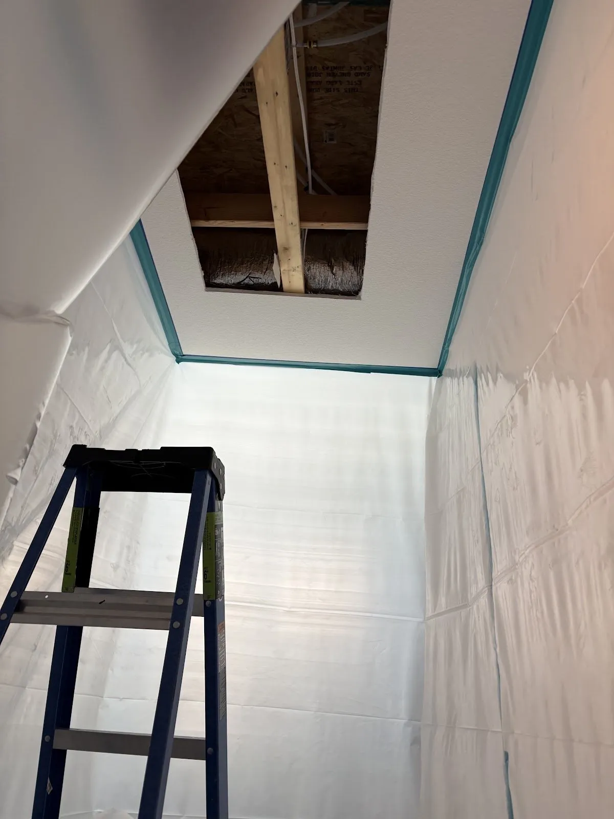 Rectangular opening cut in a lightly textured ceiling, showing wood framing and a foil-wrapped duct, with plastic sheeting taped along the walls below
