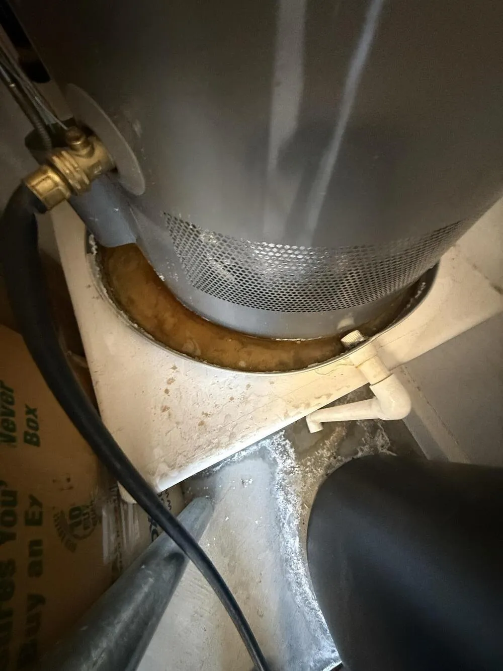 Rust-brown water pooled in a metal drain pan around the base of a gray water heater, with a brass valve at the upper left and a white plastic drain line at the edge of the pan